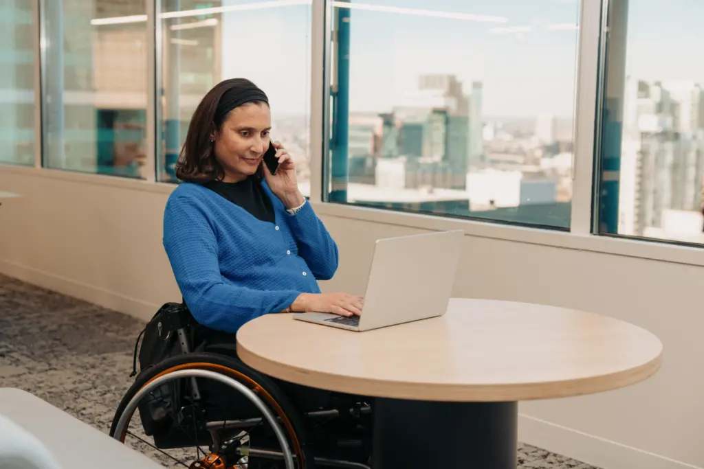 A woman takes a call in her office while working on a laptop. Her wheelchair is pulled up to the edge of a small table.